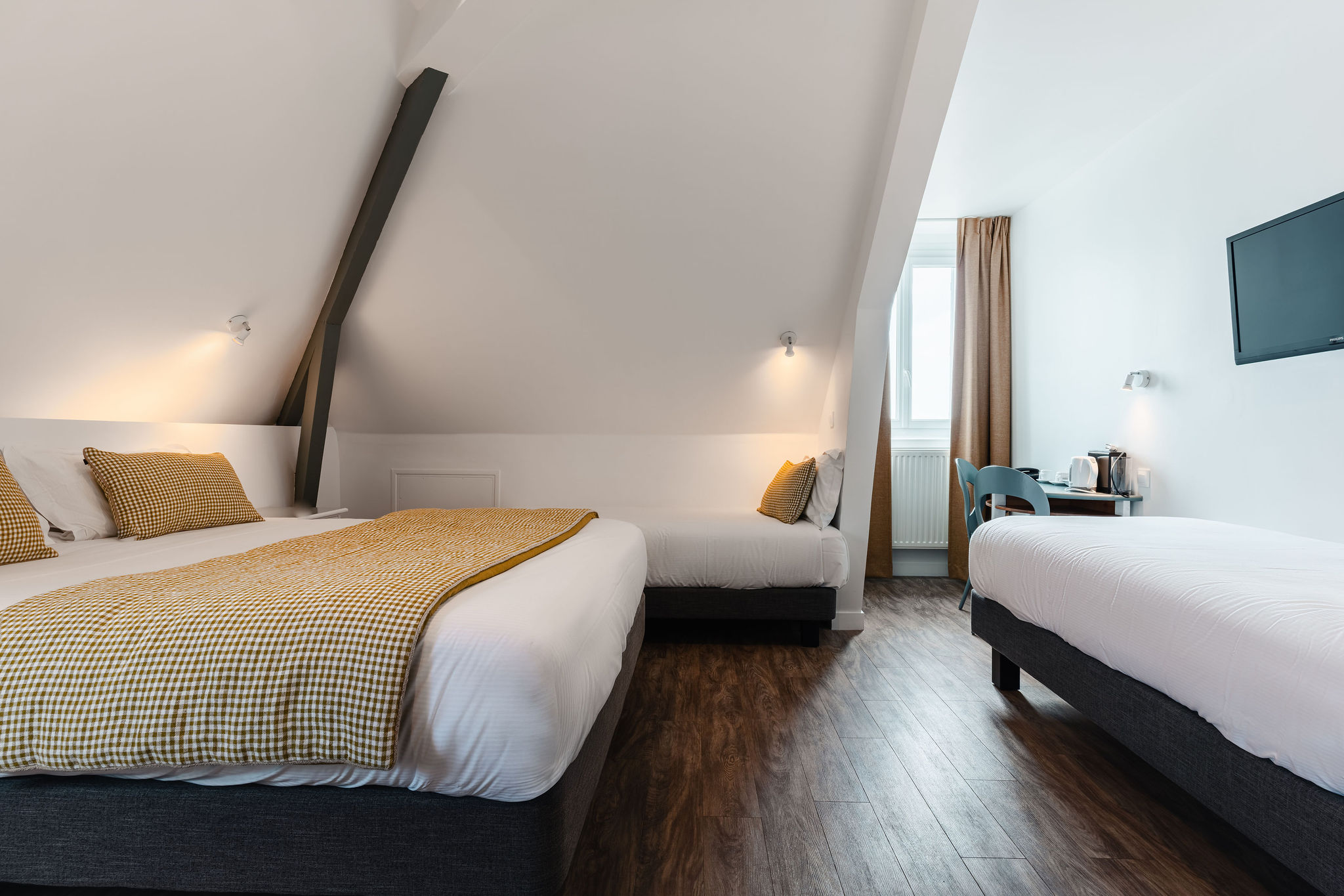 Chambre sous plafond mansardé pour 4 personnes, un lit double et deux lits simples, poutre apparente, édredon et taie d’oreillers moutarde à carreaux, murs blancs (Port-Louis, Morbihan, 56)