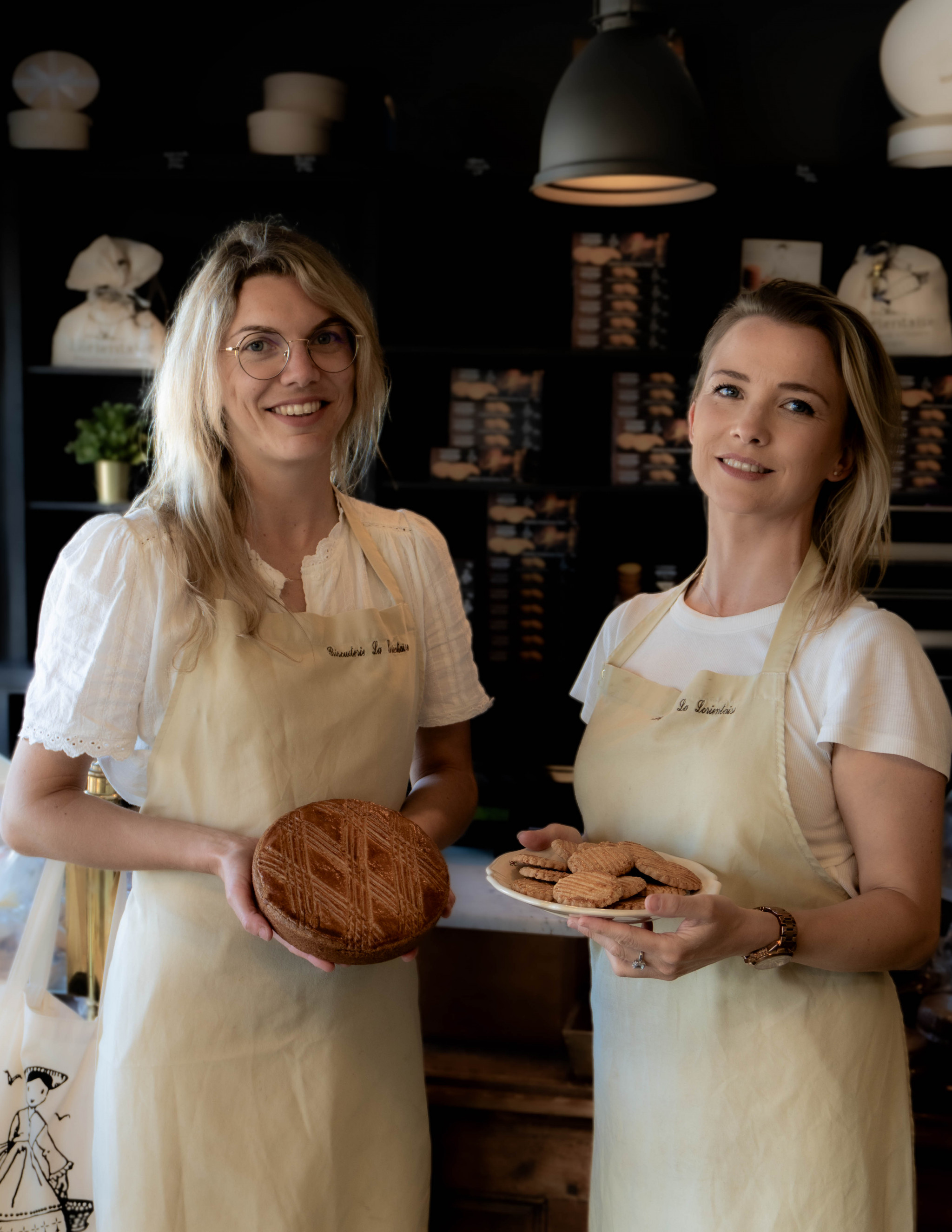 Plan serré sur les sœurs Beurel posant dans leur biscuiterie, à gauche elle présente un gâteau breton, à droite des fines galettes au beurre (Kervignac, Morbihan, 56)