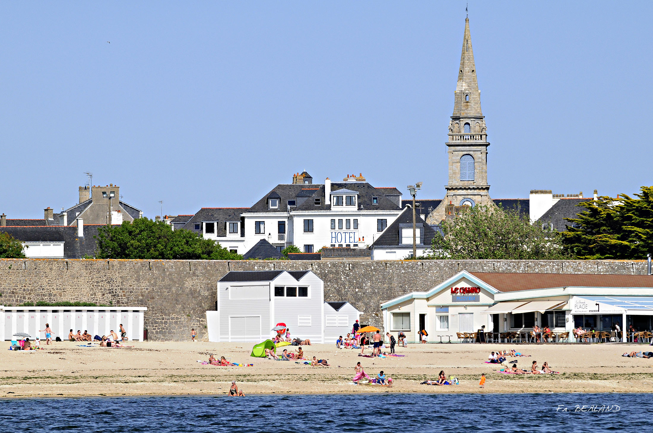 Principale – Façade avant de l’hôtel, remparts de la ville, la grande plage, son restaurant et ses cabines, clocher de l’église (Port-Louis, Morbihan, 56)