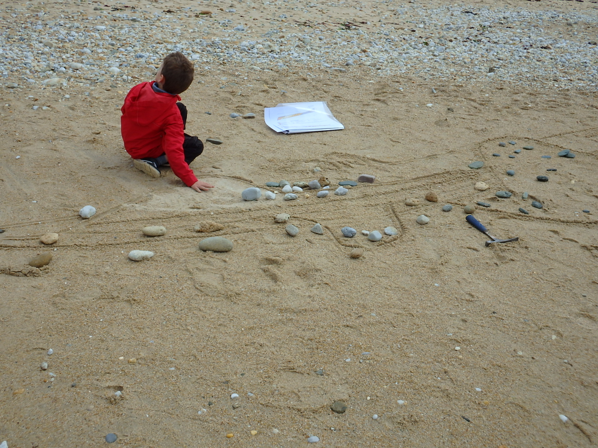 Un enfant joue sur la plage à proximité de dessins réalisés dans le sable par Samuel Piriou, le Géologue Ambulant, dans le cadre de son activité (Plouhinec, Morbihan, 56)