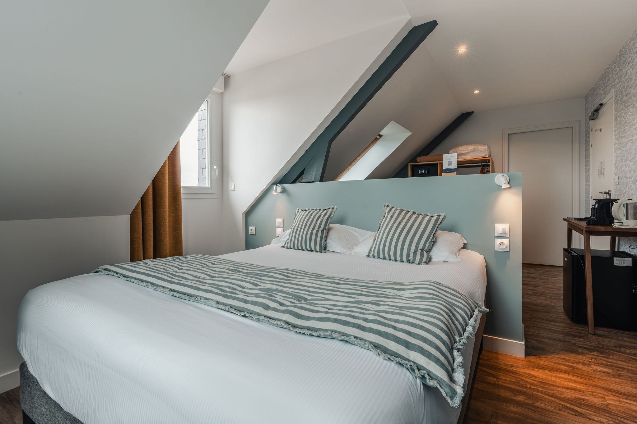 Une chambre de l’hôtel avec parquet, sous plafond mansardé, rideaux ocre épais, décoration marine rayée et tons blancs (Port-Louis, Morbihan, 56)