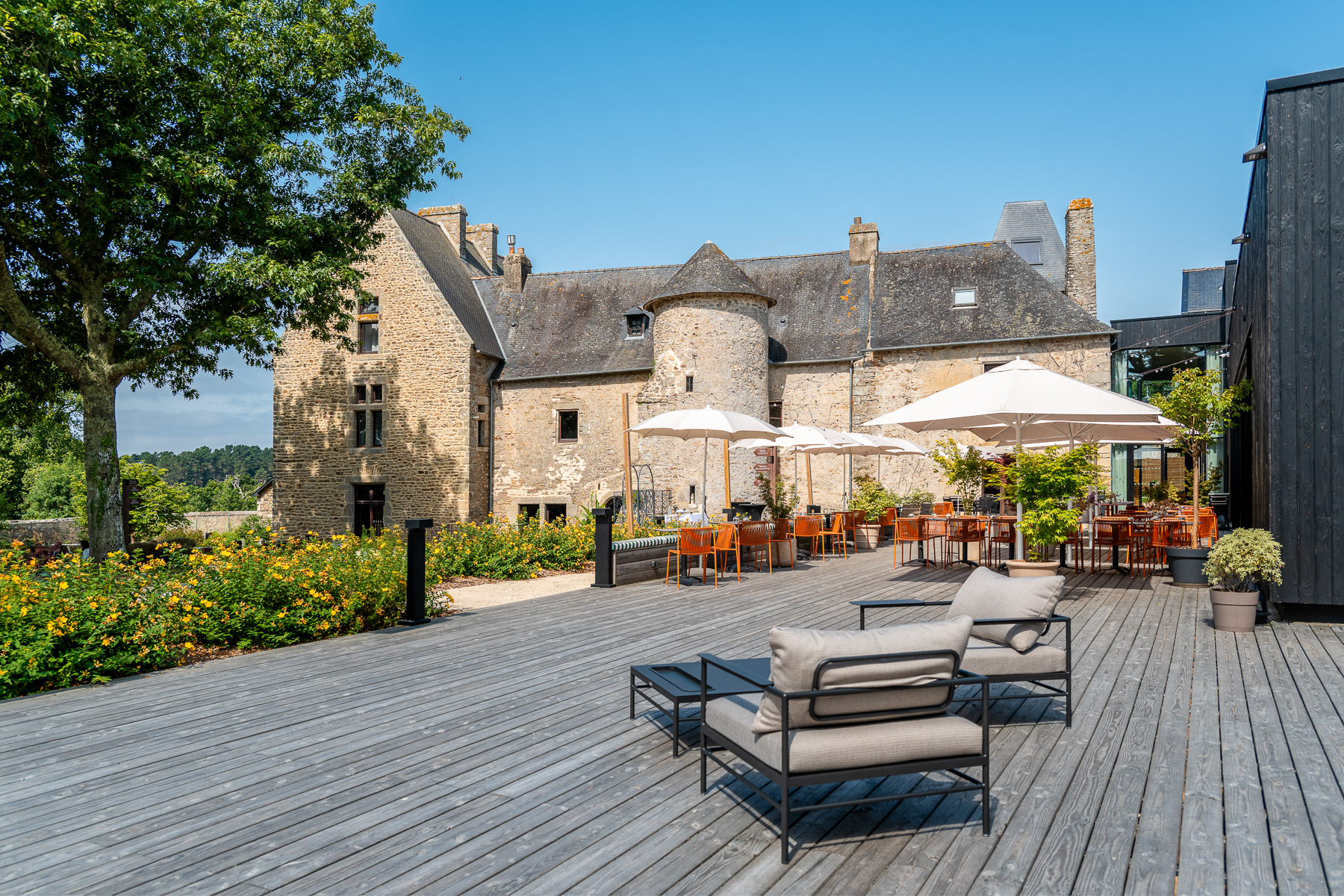 Vue sur la terrasse extérieure en bois, aménagée avec des transats, tables et parasols ainsi que sur le manoir restauré de Kerdréan (Le Bono, Morbihan, 56)