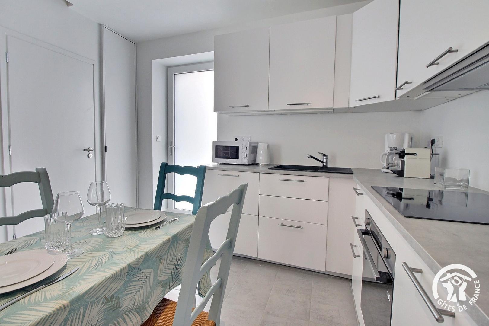 Cuisine équipée dans les tons clairs, gris/blanc avec une table dressée, vue sur une porte vitrée donnant sur l’extérieur (Sainte-Hélène, Morbihan, 56)